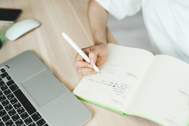 person with laptop open as they write something in notebook